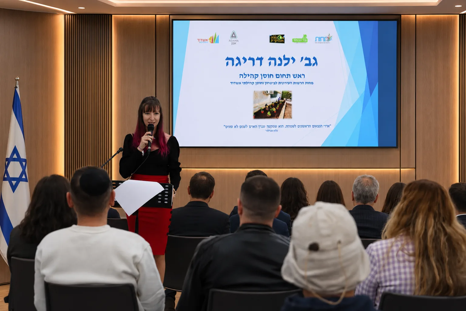 ילנה דריגה מרצה באירוע מקצועי עם מצגת על מסך גדול.